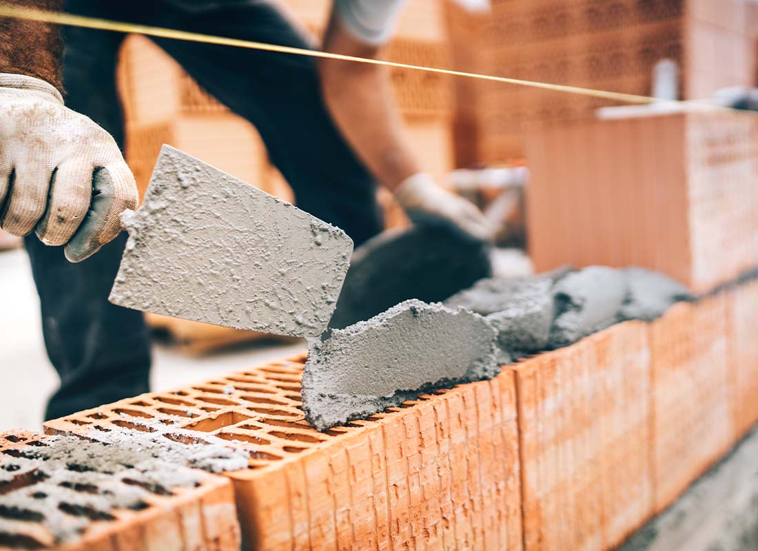 Masonry Contractor Insurance - Close-up of Construction Worker With Protective Gear and Trowel and Mortar Building Brick Walls