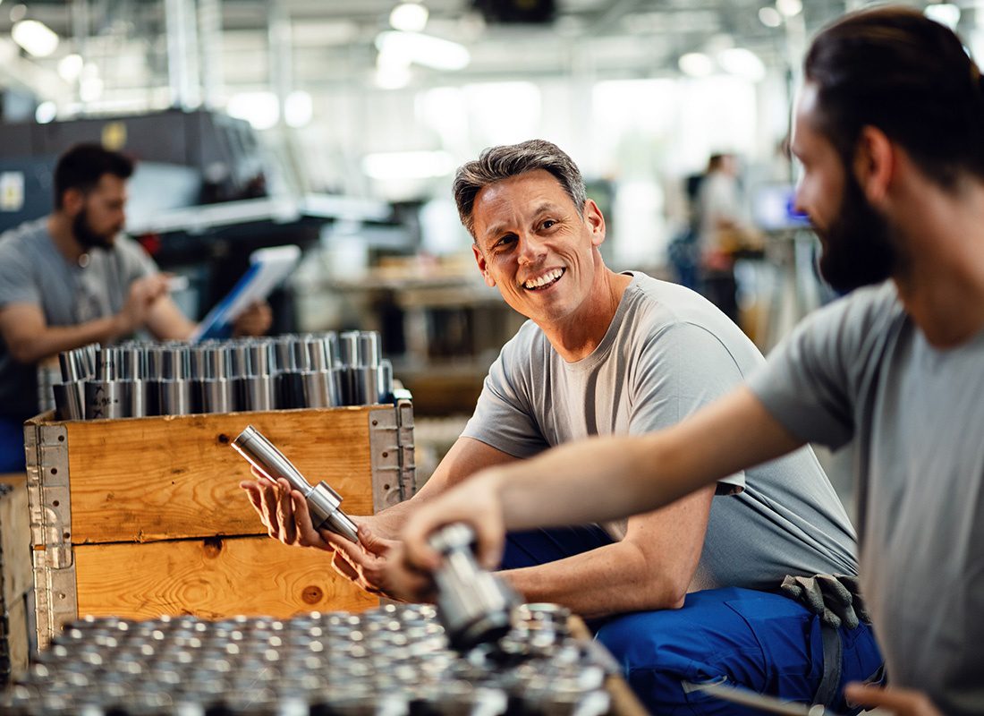 Insurance by Industry - Portrait of a Smilng Middle Aged Factory Worker Holding Steel Pipes While Looking at his Coworker Inside a Manufacturing Facility